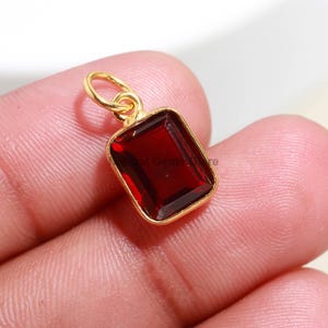Puede incluir: Un colgante rectangular de piedra preciosa rojo oscuro, con bisel y lazo dorados. El colgante se sostiene entre los dedos, mostrando la superficie pulida y facetada de la gema. El engaste dorado contrasta con el intenso color rojo.