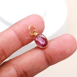 May include: A gold-colored pendant with a pink oval gemstone. The gemstone is set in a gold bezel and has a loop at the top for a chain. The pendant is held by a person's finger.