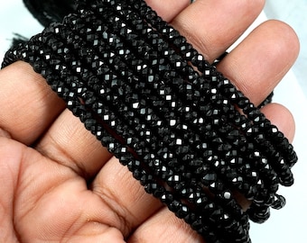 May include: A strand of black faceted gemstone beads. The beads are small and have a shiny, polished finish. The strand is coiled and held in a hand.