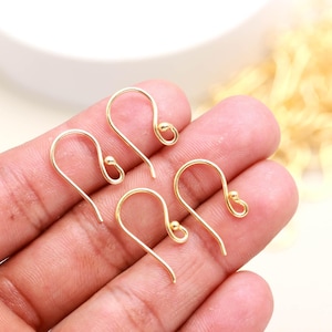Könnte beinhalten: Vier ohrhaken aus goldfarbenem metall, jeder mit einem geschwungenen drahtdesign und einem kleinen kugelakzent am ende. Diese schmuckzubehörteile haben die form von fischhaken und dienen dazu, dekorative elemente zur ohrringherstellung zu befestigen.