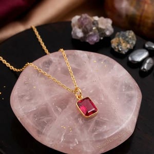 May include: A gold chain necklace featuring a rectangular, faceted, deep pink gemstone pendant set in a gold bezel. The necklace rests on a smooth, circular, pale pink rose quartz slab, with the text Natural Gems Glare visible on the surface.
