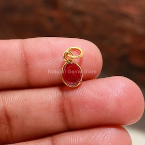 May include: A gold pendant featuring a red oval gemstone. The pendant is held on a finger.