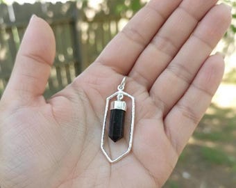 May include: A silver pendant with a black crystal gemstone. The pendant has a geometric frame and a small loop for a chain. The gemstone is a dark, opaque color and is cut into a pointed shape.