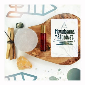 May include: A wooden tray with a white card that says "Moonbeams + Stardust" on it. There is a small bottle of red liquid, a white round stone, a piece of orange crystal, and a piece of brown wood on the tray.
