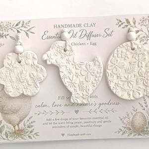 May include: A handmade clay essential oil diffuser set featuring three white textured ornaments in the shapes of a flower, a chicken, and an egg. Each piece has a floral relief pattern and a white bead on a string. The display card includes text.