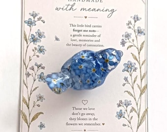 May include: A bird-shaped resin ornament containing embedded blue forget-me-not flowers, mounted on a rectangular card featuring floral illustrations, a small bird graphic, and the text: Handmade with meaning, This little bird carries forget me nots, Those we love don't go away, they bloom in the flowers we remember, thank you FOR SUPPORTING HANDMADE.