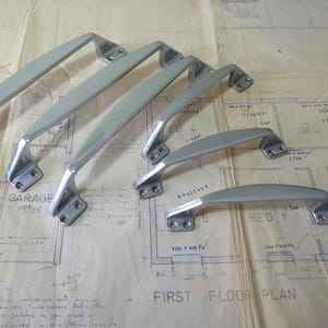 May include: Several silver-coloured metal door or cabinet handles. The handles have a curved design and are mounted on rectangular bases with screw holes. The items are arranged on a vintage blueprint.