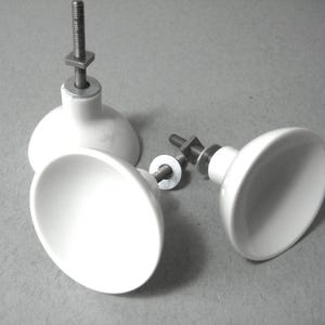 May include: Three white ceramic cabinet knobs with metal fixings. The knobs have a rounded, oval shape and are attached to threaded rods with nuts and washers. The knobs are on a grey surface.