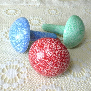 May include: Three mushroom-shaped decorative objects with a speckled finish. One object is red with white speckles, one is blue with white speckles, and one is green with white speckles. Each piece features a rounded cap and a cylindrical stem.