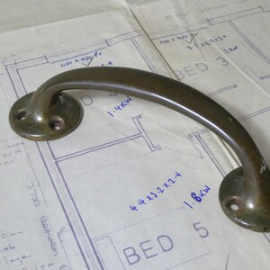 May include: A bronze-coloured door handle with a curved bar and circular mounting plates. The handle rests on a blueprint with handwritten notes and measurements, including "BED 5" and "1.8kW".