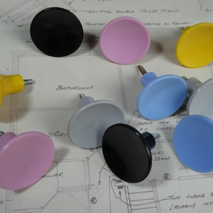 May include: A collection of nine round cabinet knobs in various solid colours including black, pink, yellow, light blue, and grey. Each knob features a smooth, circular face and a metal screw protruding from the back for installation purposes.