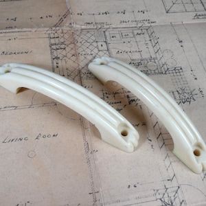 May include: Two off-white, curved cabinet handles featuring a triple-ribbed design. Each handle has two circular mounting holes at the ends, with a smooth, glossy finish. The handles are shaped with a slight arch and have rounded, textured tips.