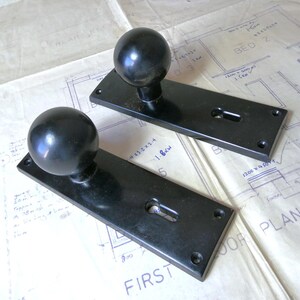May include: Two black door handles with spherical knobs mounted on rectangular backplates. Each backplate features a keyhole cutout and four screw holes at the corners. The hardware has a smooth, dark finish and a simple, functional design.