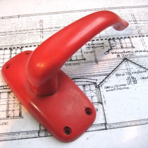 May include: A red plastic door handle with a rounded handle and a rectangular base. The handle is on a white surface with black lines.