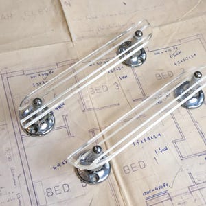 May include: Two clear acrylic drawer pulls with polished chrome metal bases. Each handle features a long, rectangular, transparent bar supported by two circular chrome mounting plates with visible screw holes at each end of the hardware.