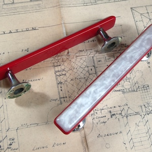 May include: Two rectangular cabinet pulls with metallic mounting hardware. One pull features a solid glossy red finish, while the other has a red frame with a white marbled inlay. Both handles are positioned diagonally across a technical blueprint.