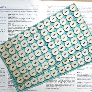 May include: Two cards of cream-coloured, round buttons with two holes each, arranged in a grid pattern. The buttons are on a pale blue grid, with knitting instructions in the background.