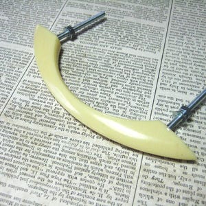 May include: A cream-coloured plastic cabinet door handle with a curved design and silver metal screws.