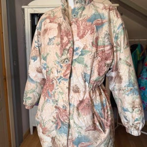 Puede incluir: Chaqueta vintage con estampado floral, cuello alto y cierre de botones. La chaqueta presenta una cintura con cordón y un estampado de flores rosas, azules y verdes sobre fondo crema. Mangas largas y dos bolsillos delanteros.