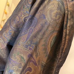 Puede incluir: Primer plano de una prenda con un estampado de cachemira en tonos morado, dorado y verde azulado. La tela tiene brillo y el diseño es intrincado. La prenda parece ser una chaqueta o abrigo, con costuras visibles y un bolsillo.