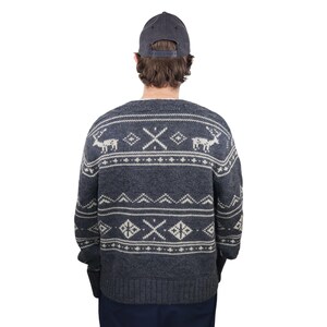May include: A gray sweater with a white reindeer and geometric pattern. The sweater has long sleeves and a crew neck. The person is wearing a dark blue baseball cap.
