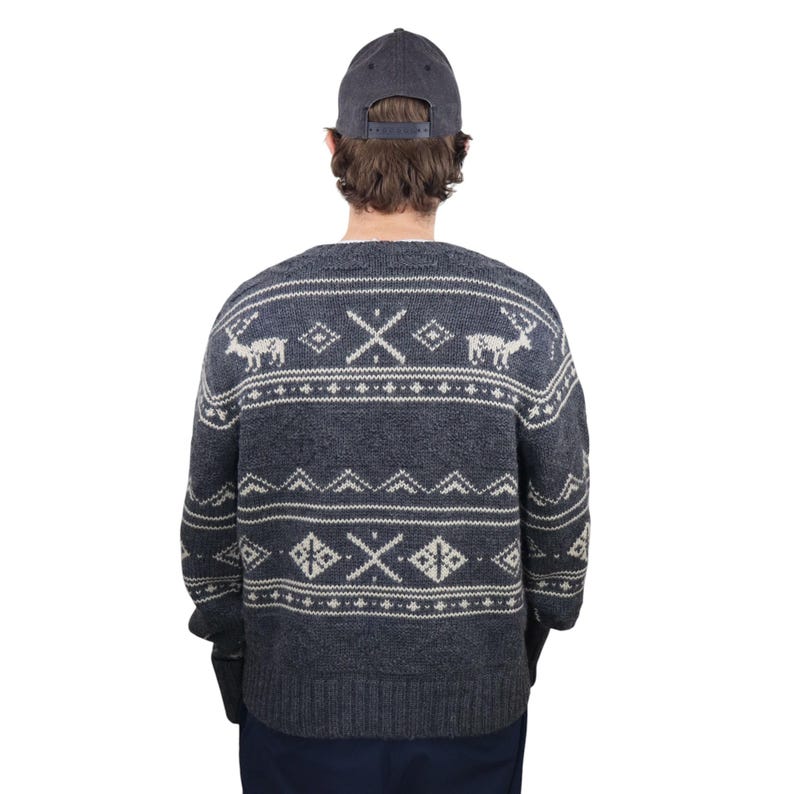 May include: A gray sweater with a white reindeer and geometric pattern. The sweater has long sleeves and a crew neck. The person is wearing a dark blue baseball cap.
