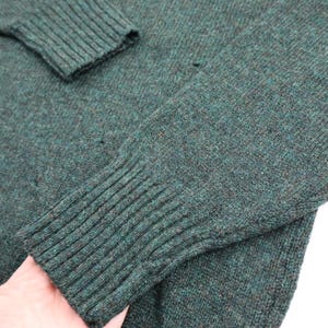 Pu&ograve; includere: Primo piano di un maglione verde scuro. I polsini e lo scollo a coste sono visibili, insieme al tessuto a maglia strutturato. Il maglione sembra essere fatto di un materiale morbido e caldo, perfetto per la stagione fredda. Il colore &egrave; un verde intenso e smorzato.