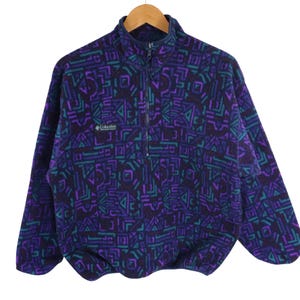 以下が含まれることがあります： 紫、ティール、黒の幾何学模様が特徴的な、ヴィンテージのコロンビア製フリースプルオーバー。ハーフジップ開閉、スタンドカラー、胸にコロンビアのロゴ入り。木製のハンガーに掛けられており、レトロなデザインが際立っています。