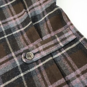 K&ouml;nnte beinhalten: Nahaufnahme eines karierten Stoffes mit Tasche und Knopf. Das Muster umfasst Braunt&ouml;ne, Grau und Lavendel, wobei wei&szlig;e Linien das Karomuster bilden. Der Knopf ist klein, rund und hat zwei L&ouml;cher.