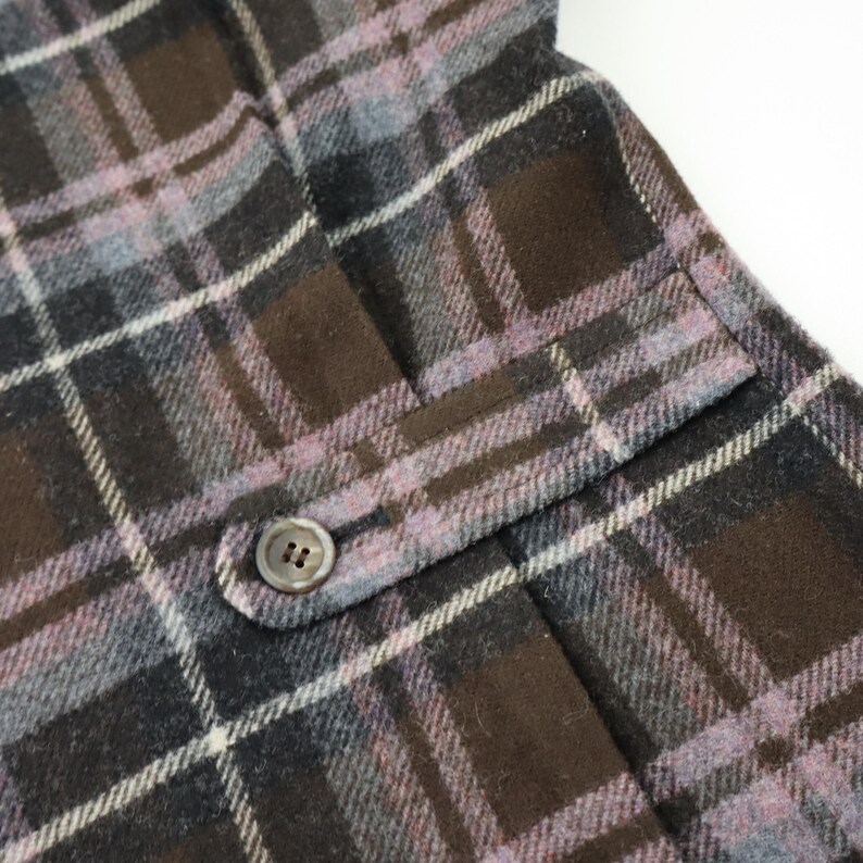 K&ouml;nnte beinhalten: Nahaufnahme eines karierten Stoffes mit Tasche und Knopf. Das Muster umfasst Braunt&ouml;ne, Grau und Lavendel, wobei wei&szlig;e Linien das Karomuster bilden. Der Knopf ist klein, rund und hat zwei L&ouml;cher.