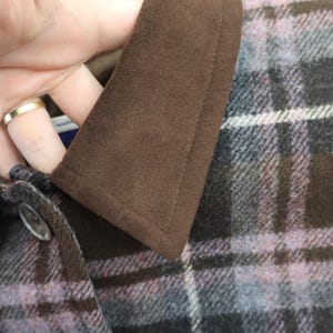 K&ouml;nnte beinhalten: Nahaufnahme eines braunen Wildleder-Kragens auf einer karierten Jacke. Die Jacke hat einen Knopfverschluss und ein Muster aus schwarzen, lila und wei&szlig;en Karos. Der Kragen ist dunkelbraun und hat eine weiche Textur.