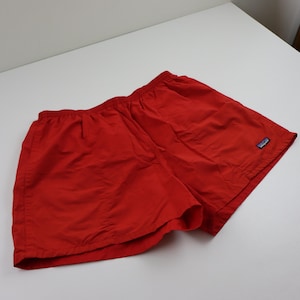 May include: Bright red shorts with an elastic waistband. The shorts have a small blue and white logo patch on the lower right side. The shorts are made of a lightweight material.