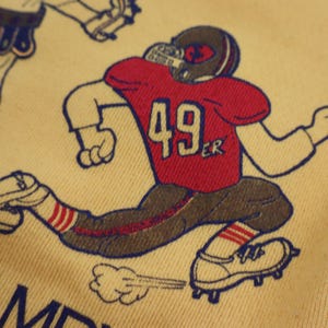 Puede incluir: Una camiseta amarilla con una ilustraci&oacute;n de dibujos animados de un jugador de f&uacute;tbol americano que lleva una camiseta roja con el n&uacute;mero 49 y la palabra "ER" impresa. El jugador corre con un bal&oacute;n de f&uacute;tbol y una nube de polvo detr&aacute;s de &eacute;l.