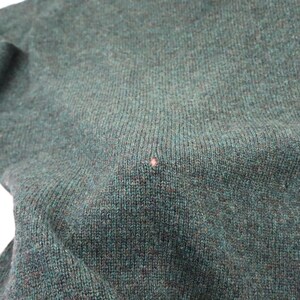 Pu&ograve; includere: Primo piano di un maglione di maglia verde scuro. Il tessuto ha un aspetto strutturato con una piccola macchia rosa chiaro. Il colore del maglione &egrave; un verde intenso e smorzato, con sottili variazioni di tono.