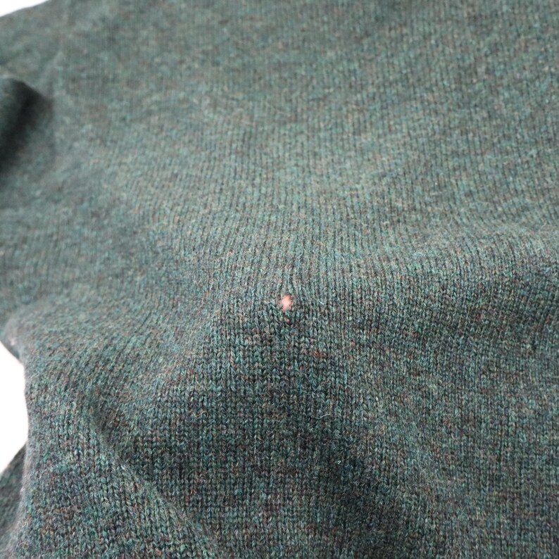 Pu&ograve; includere: Primo piano di un maglione di maglia verde scuro. Il tessuto ha un aspetto strutturato con una piccola macchia rosa chiaro. Il colore del maglione &egrave; un verde intenso e smorzato, con sottili variazioni di tono.