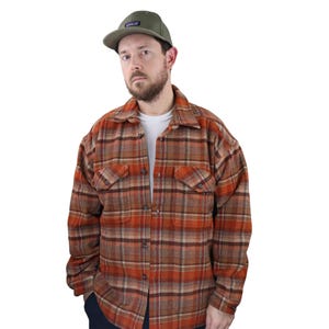 Puede incluir: Una persona que lleva una gorra de béisbol verde oliva y una camisa de franela a cuadros en tonos naranja, marrón y beige. La camisa tiene dos bolsillos en el pecho y una tapeta abotonada. La persona lleva una camiseta blanca debajo.