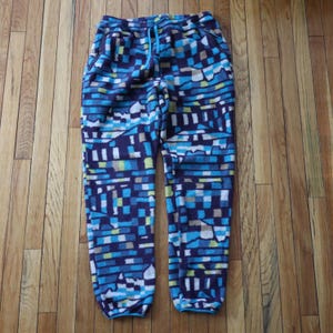 May include: Fleece trousers with a blue, white, and yellow geometric pattern, drawstring waist, and two zipped pockets. The soft, warm material trousers are approximately 102 cm long.