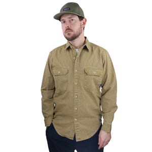 May include: A tan button-down shirt with two chest pockets and a green baseball cap. The cap has a small rectangular patch on the front. The shirt is buttoned up and has long sleeves. The person is wearing dark blue pants.