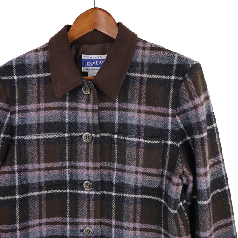 K&ouml;nnte beinhalten: Pendleton-Jacke mit braunem, schwarzem und lavendelfarbenem Karomuster, mit braunem Kragen und Knopfverschluss. Die Jacke h&auml;ngt an einem Holzb&uuml;gel. Das Pendleton-Etikett ist sichtbar.