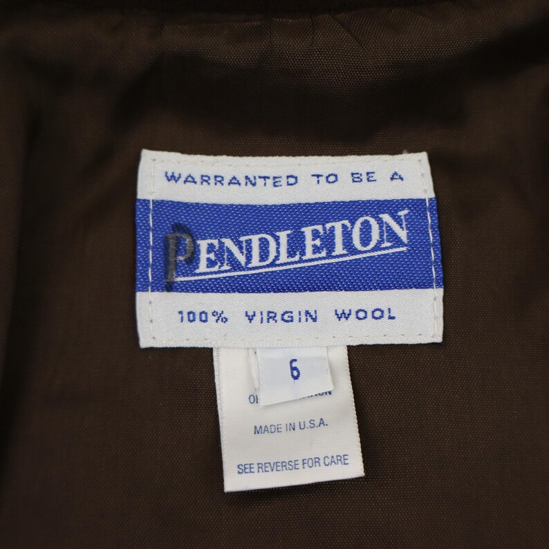 K&ouml;nnte beinhalten: Nahaufnahme eines Pendleton-Etiketts mit dem Markennamen in Wei&szlig; auf blauem Hintergrund. Das Etikett enth&auml;lt auch die Aufschrift "100% Virgin Wool" und "Made in U.S.A.". Das Gr&ouml;&szlig;enetikett zeigt "6".