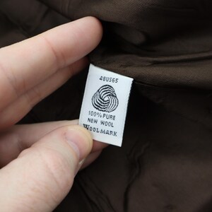 K&ouml;nnte beinhalten: Nahaufnahme eines wei&szlig;en Etiketts mit der Aufschrift "48US65", einem Wollsiegel-Symbol und den Worten "100% Pure New Wool Woolmark". Das Etikett wird vor einem braunen Stoffhintergrund gehalten.
