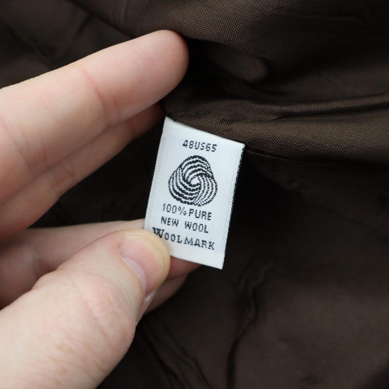 K&ouml;nnte beinhalten: Nahaufnahme eines wei&szlig;en Etiketts mit der Aufschrift "48US65", einem Wollsiegel-Symbol und den Worten "100% Pure New Wool Woolmark". Das Etikett wird vor einem braunen Stoffhintergrund gehalten.