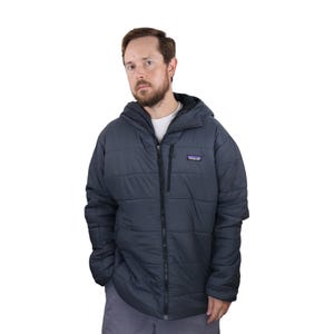 May include: A dark blue quilted jacket with a hood and a zipper closure. The jacket has a small logo patch on the chest and is worn by a person. The jacket appears to be made of a lightweight material.