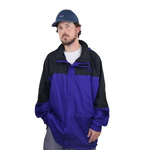 Puede incluir: Una gorra de béisbol azul con logotipo y una chaqueta de dos tonos con una sección superior negra y una sección inferior morada. La chaqueta tiene cuello alto, cierre de cremallera y mangas largas, sobre una camiseta blanca lisa.