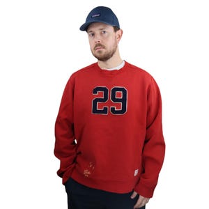 Puede incluir: Sudadera roja con un número 29 azul marino en la parte delantera. La sudadera tiene cuello redondo y mangas largas.
