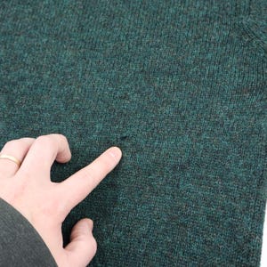 Pu&ograve; includere: Primo piano di un maglione verde scuro con un piccolo foro. Il maglione ha un motivo a maglia strutturato. Una mano con un anello d'oro indica il foro nel maglione.