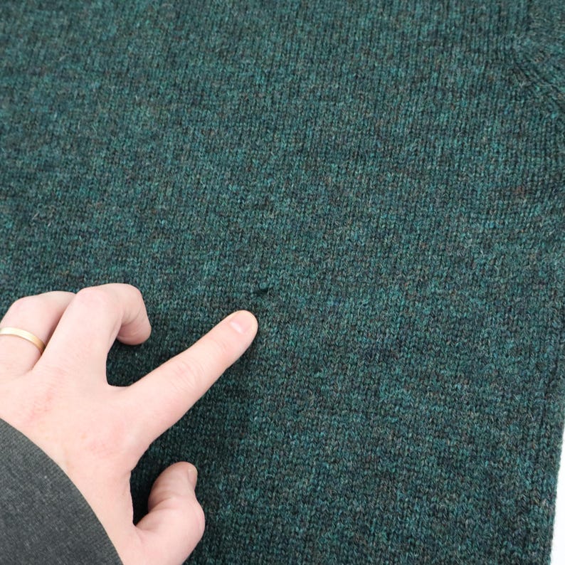 Pu&ograve; includere: Primo piano di un maglione verde scuro con un piccolo foro. Il maglione ha un motivo a maglia strutturato. Una mano con un anello d'oro indica il foro nel maglione.