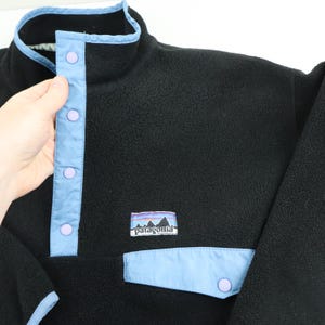 Może przedstawiać: Czarny polar z jasnoniebieskimi wykończeniami i akcentami. Posiada zapięcie na guziki i małą kieszeń z jasnoniebieską klapką. Logo Patagonia jest widoczne na piersi.