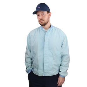 May include: A light blue bomber jacket with a matching collar and cuffs. The jacket has a button-up front and two pockets. A navy blue baseball cap with a small logo is worn on the head.