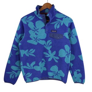 Puede incluir: Un jersey de forro polar azul con un estampado floral verde. El jersey tiene un cierre de botones a presión y un pequeño bolsillo en el pecho izquierdo. El logotipo de Patagonia es visible en el bolsillo.
